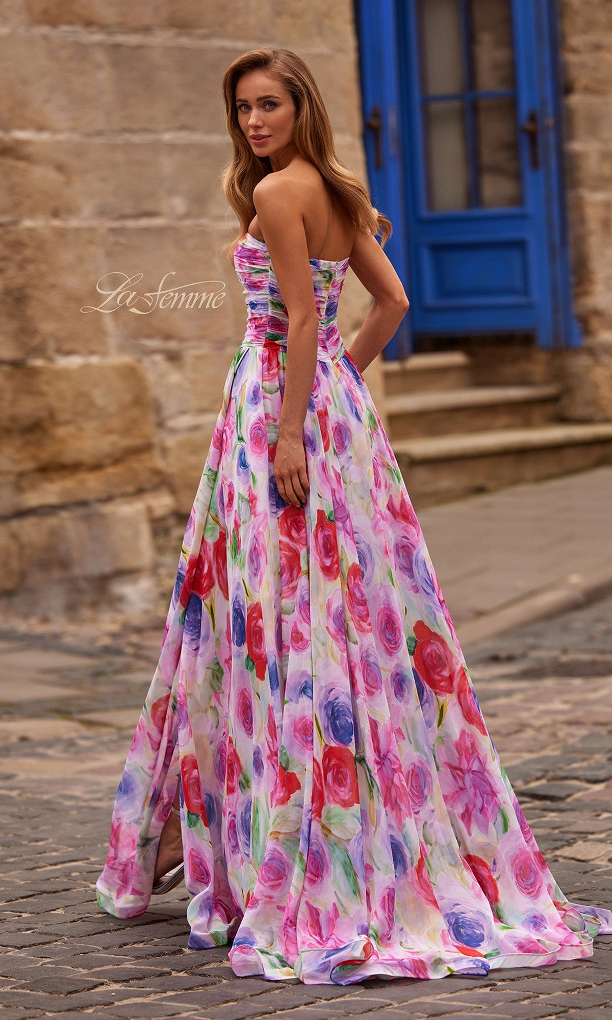 A woman in the La Femme 33487 long prom dress with a strapless, ruched, multi-color floral bodice stands on a cobblestone street by a blue door and stone building, glancing over her shoulder.